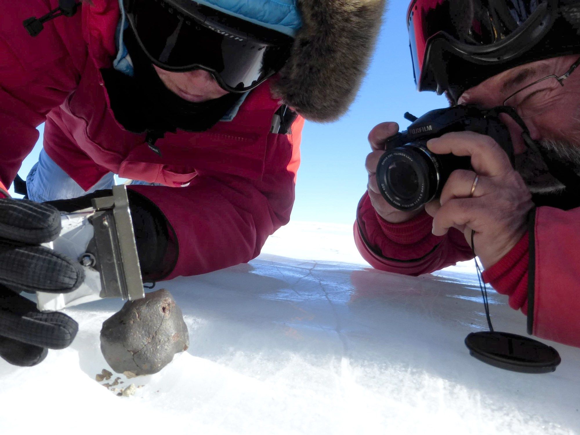 Scientists in heavy red coats and eye goggles photograph a meteorite in the snow.
