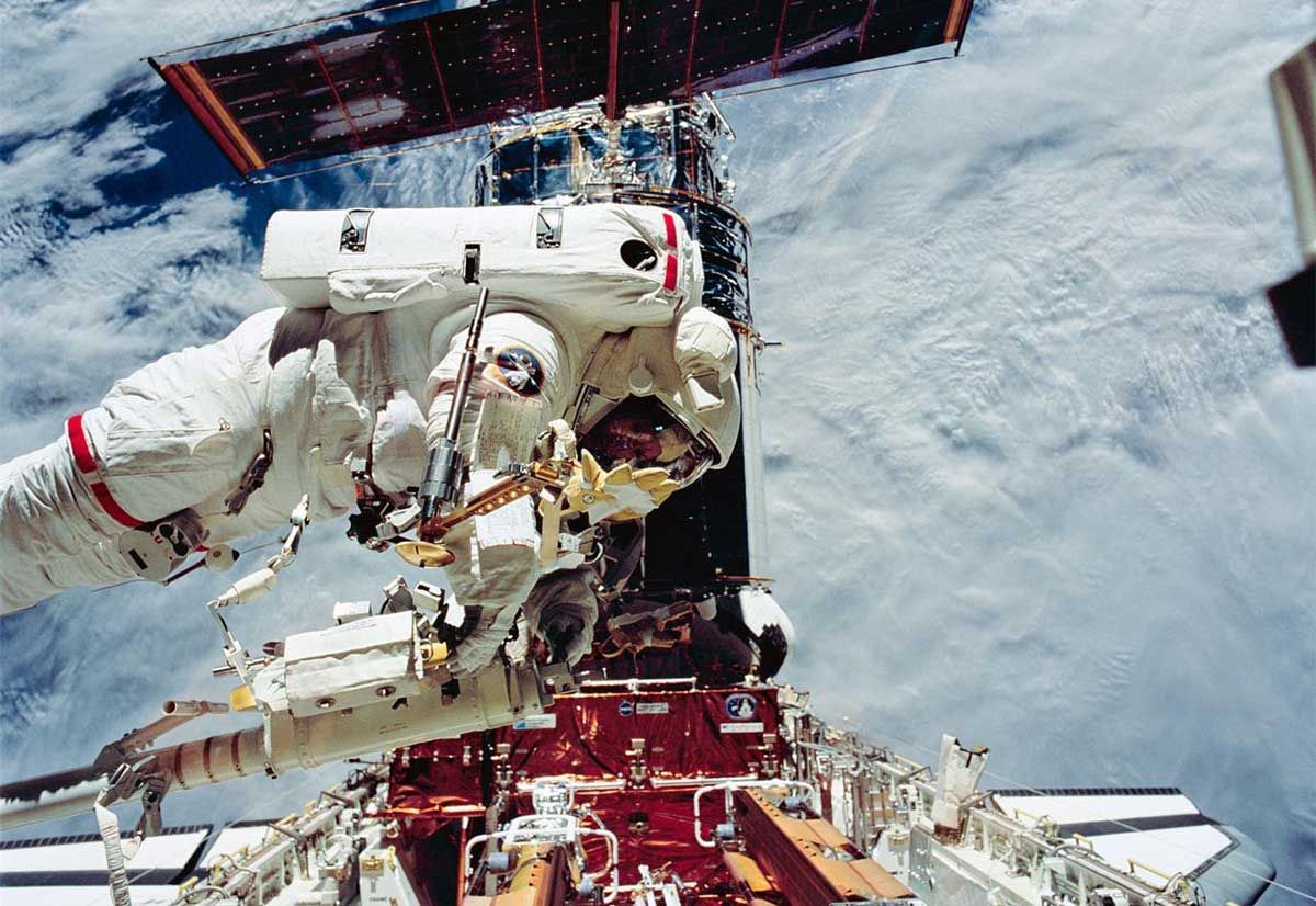 Image showing astronaut floating above the blue Earth, in the background you see Hubble with one of its solar arrays. The Shuttle is also visible at the bottom of the image.