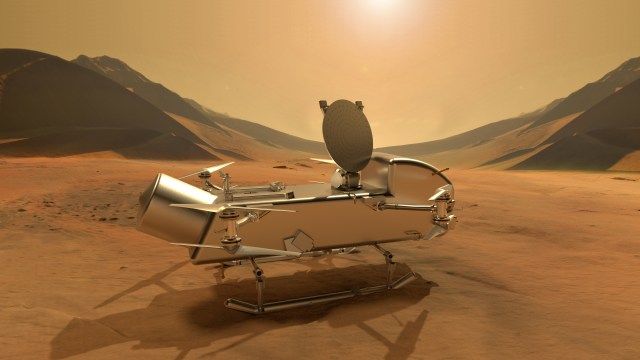 A shiny metal spacecraft is shown sitting on a brown, sandy surface. Sand dunes are shown rising on the left and right in the background.