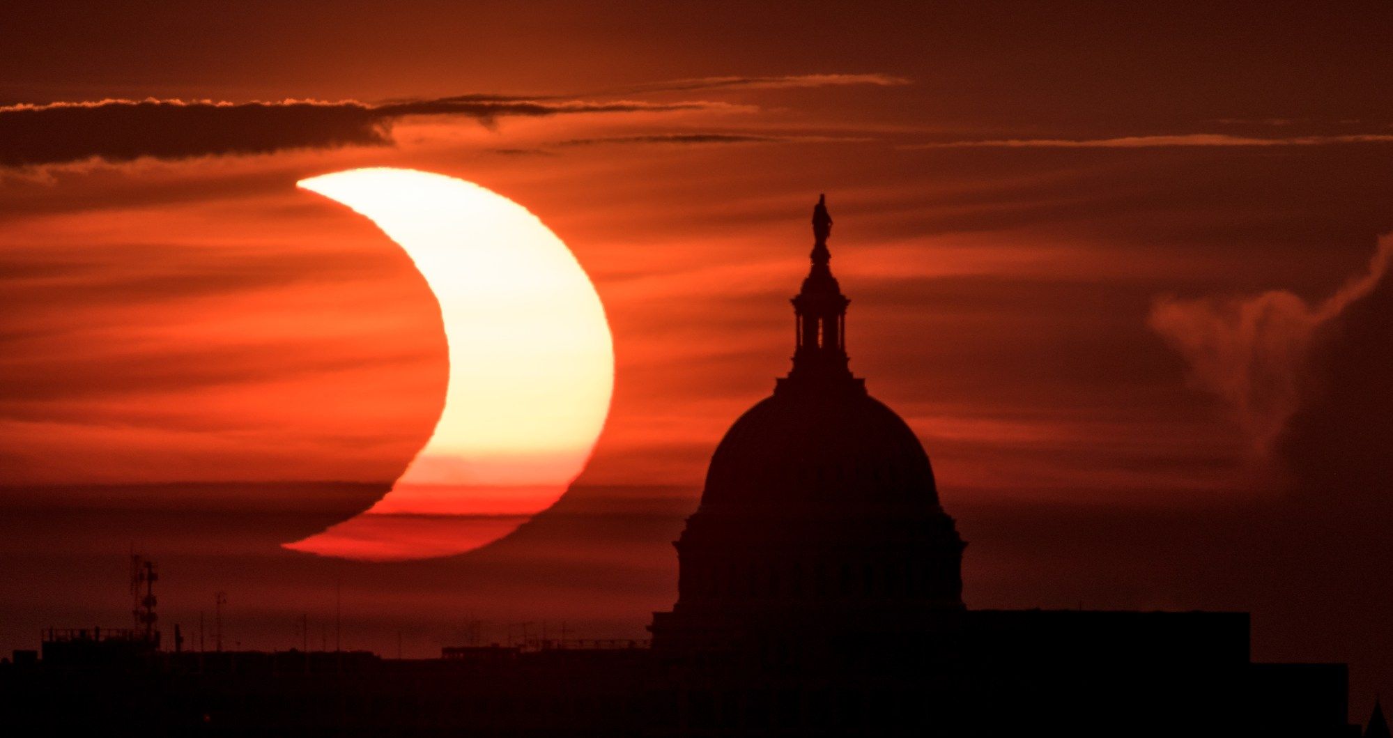 A crescent Sun against a dark orange and red background. It's to the left of the top of the U.S. Capitol Building, shown in silhouette.