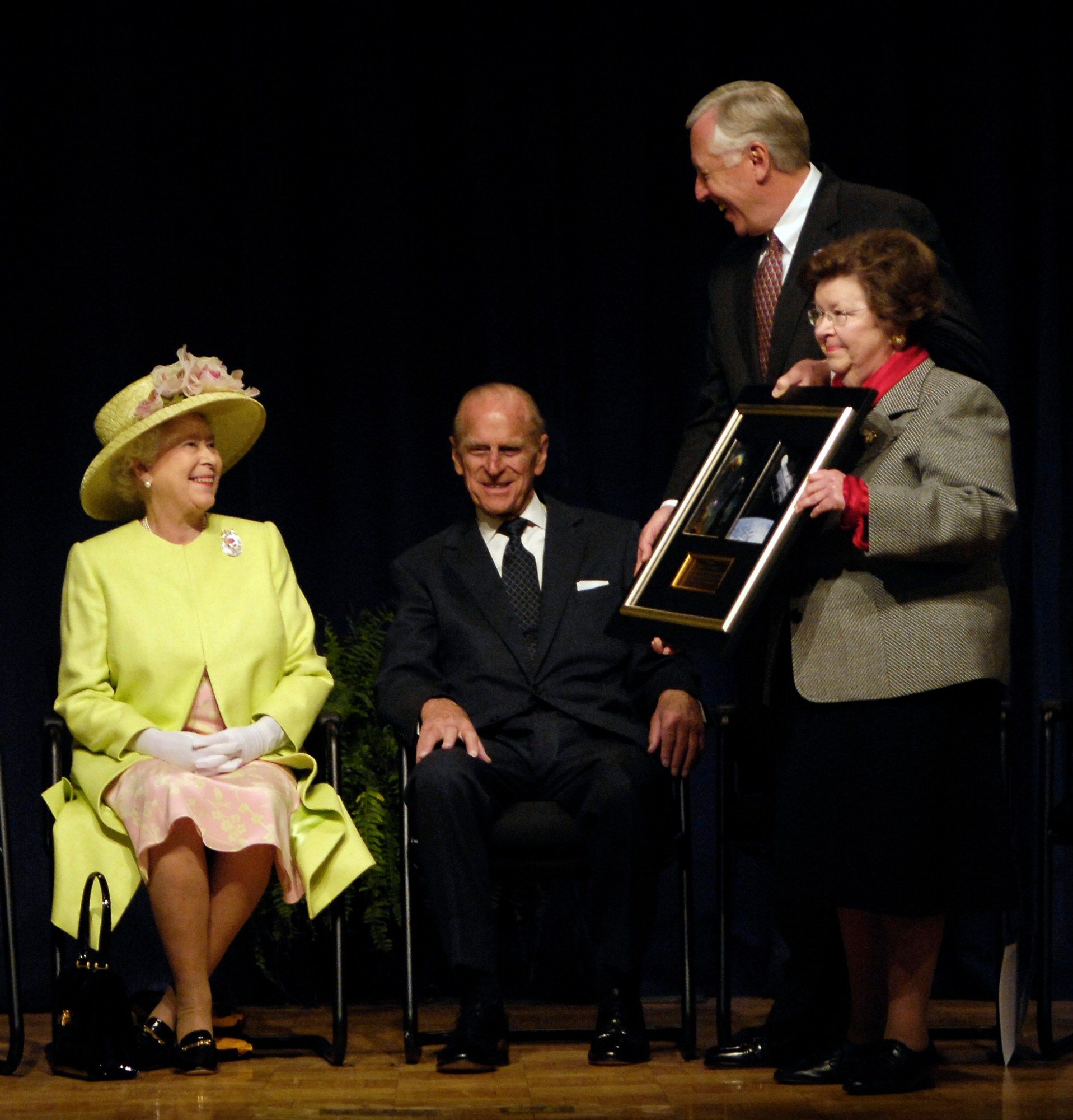 Queen Elizabeth of England, wearing a yellow coat and yellow hat with pink flowers and seated next to Prince Phillip in a dark suit, is presented with a framed Hubble image featuring photographs of a cosmic object and the telescope by Sen. Barbara Mikulski and Rep. Steny Hoyer.