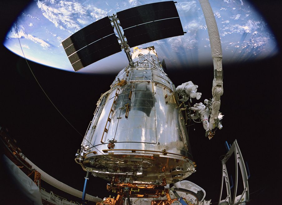 An astonaut works on the Hubble Space Telescope above the Earth as seen from a fish eye camera.