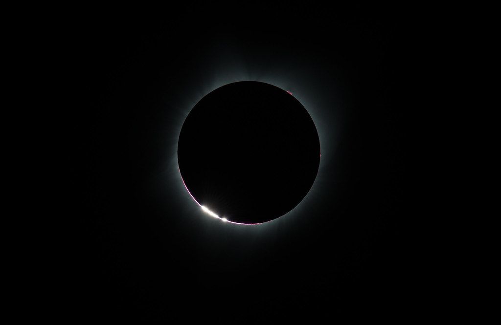 A close-up photograph of a solar eclipse, showing a bright cluster of orbs from the Sun on the lower left edge where sunlight peaks around the Moon.