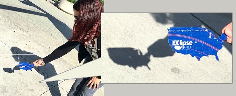 A woman holds a pinhole projector in her hand. The project is shaped like the U.S. and has a small hole in the center. Behind the projector on the ground, there is the shadow of the projector. In the middle, a small, crescent of light appears. This is the shape of the Sun.