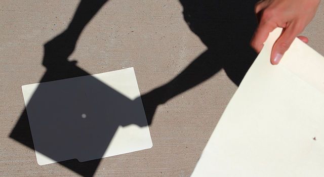 On the right, a white paper with a small hole poked in the middle takes up the image. A hand holds the paper. On the left, a shadow of the paper is on the ground. In the middle, a small circle of light shines through -- representing the Sun.