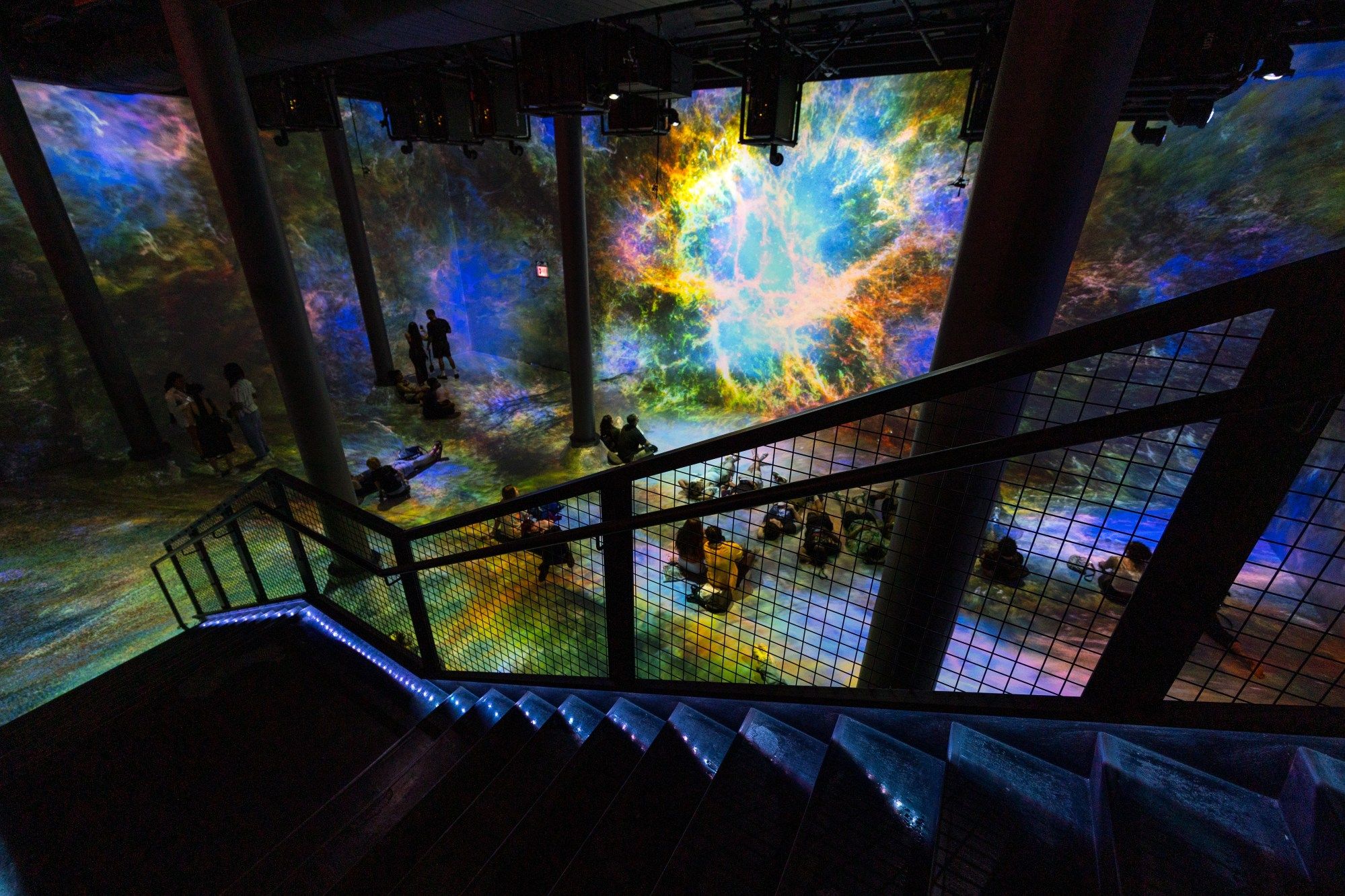 People sit and stand at an art exhibit in a dark room surrounded by tall projected Hubble images.