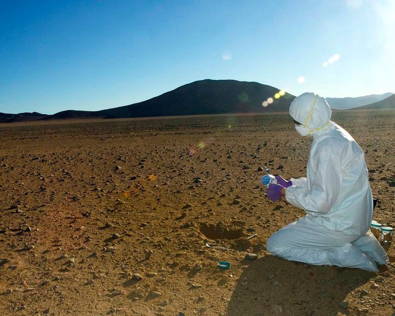 A researcher in a clean suit kneels in the desert, collecting samples.