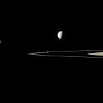 Against a black background, five golden-gray moons of varying sizes are scattered along a horizontal band through the image. Sunlight illuminates them from the left, and their right sides are in shadow. The largest, at far right, takes up the middle third of the photo, fully sunlit, with its dark half cut off by the right edge of the frame. The rings of Saturn, seen nearly edge-on, pierce the image like a knife blade from center-right, emerging through the largest moon, and with the smallest moon resting atop the blade like a breadcrumb.