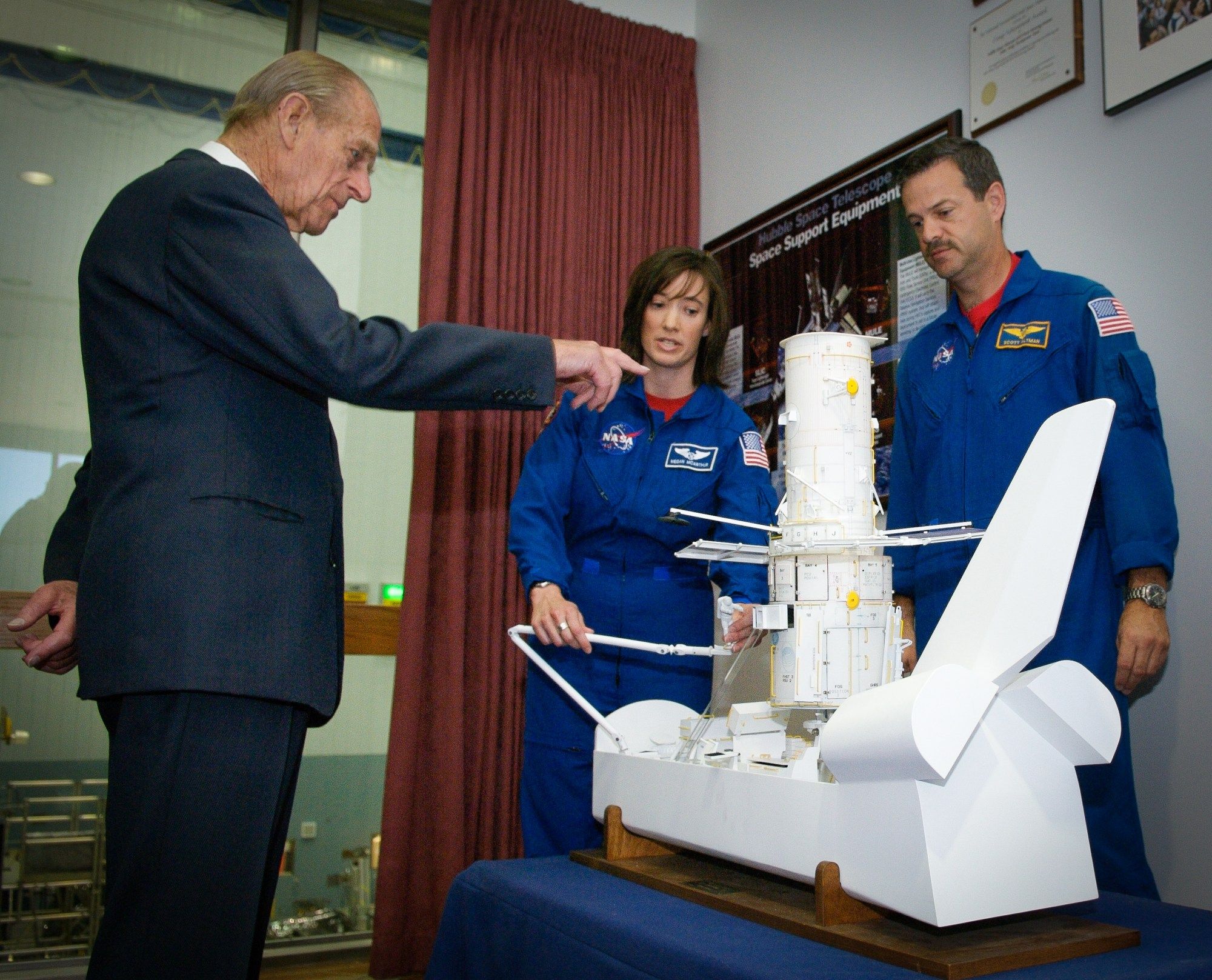 Two astronauts in blue uniforms stand behind a simple model of the space shuttle that has a detailed model of the Hubble Space Telescope in its cargo bay. One holds the shuttle's model robotic arm.Prince Phillip stands to the left and points at the Hubble model.