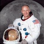 Headshot of man in a white space suit holding a helmet in front of a backdrop photo of the moon