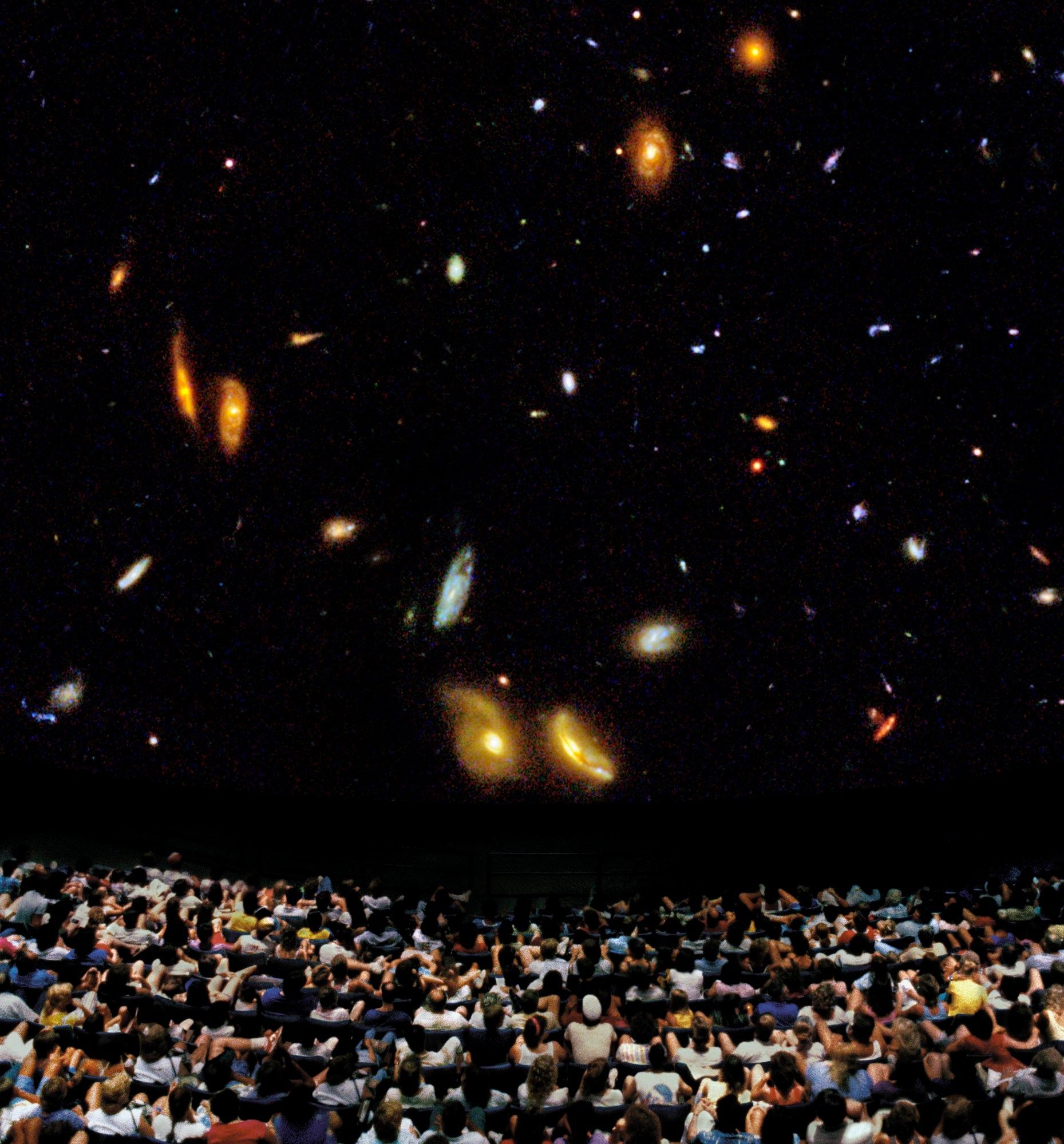 An audience in a theater watches a large format film showing galaxies.