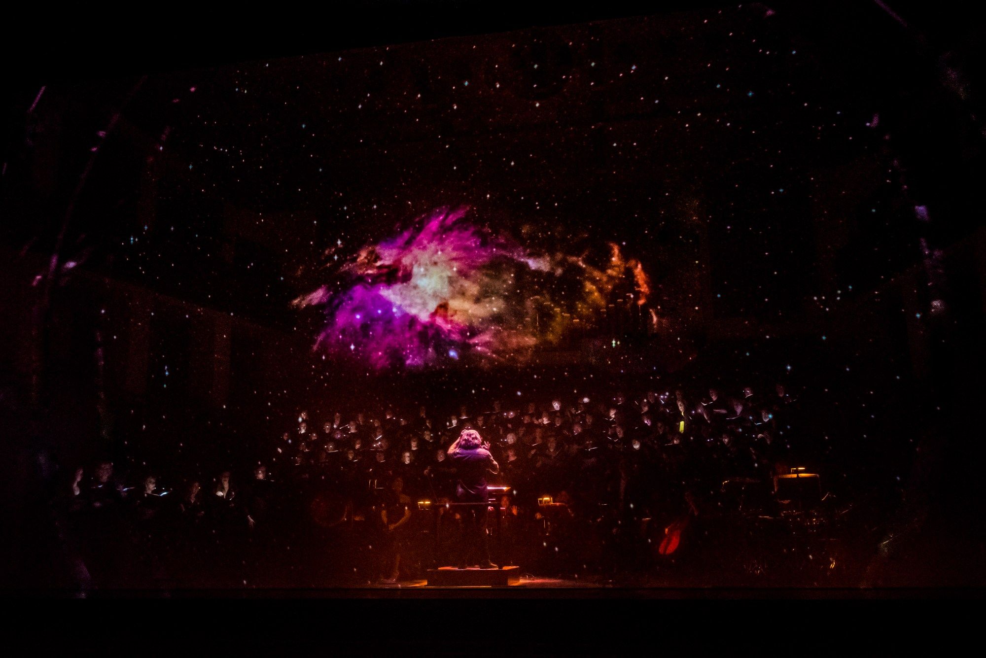 A conductor leads a choir on a darkened stage with a purple and orange cosmic nebula image hovering over the group and bathing then in its glow.