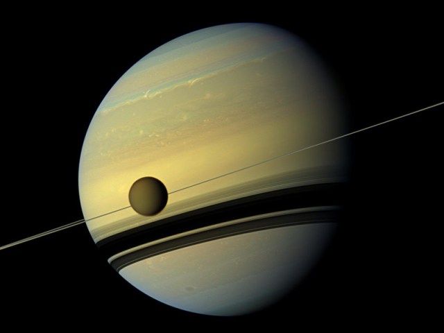 A global view of Saturn showing its yellow-brown structure. Part of its right section is in shadow. Two large bands of black spread across its lower half. A dark yellow-brown circle hovers in front of Saturn at the middle-left.