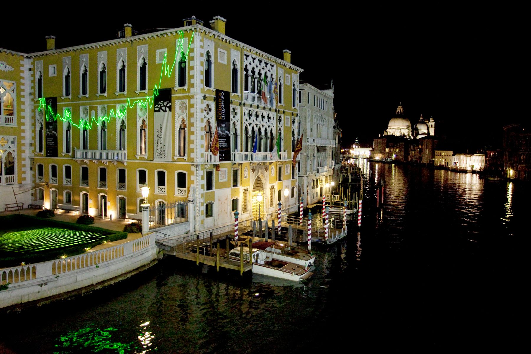 A jagged green line of laser light, resembling an EKG, plays across the face of a yellow and white building on the Venice canals at night.