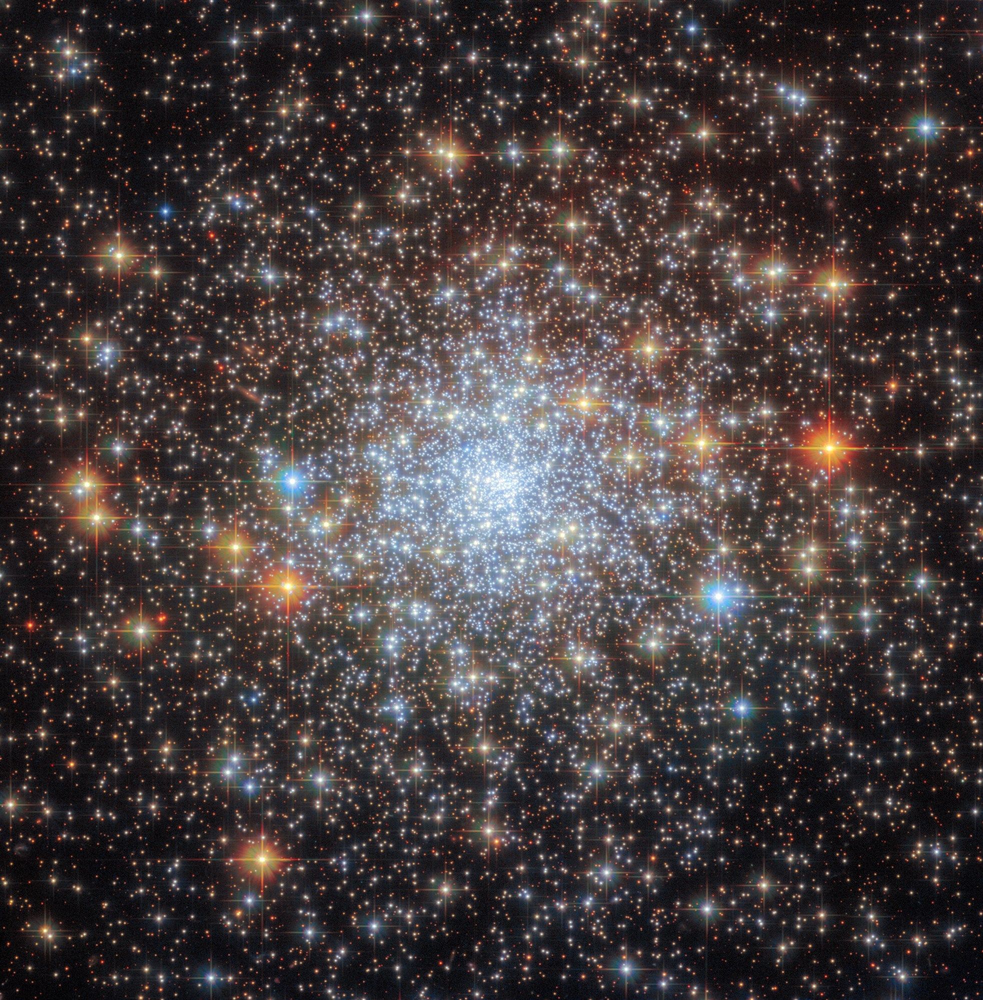 A dense spherical cluster of stars. The stars merge into a bright core in the center, and spread out to the edges gradually, giving way to an empty, dark background. Most of the stars are small points of light. A few stars with cross-shaped diffraction spikes appear larger and stand out in front.