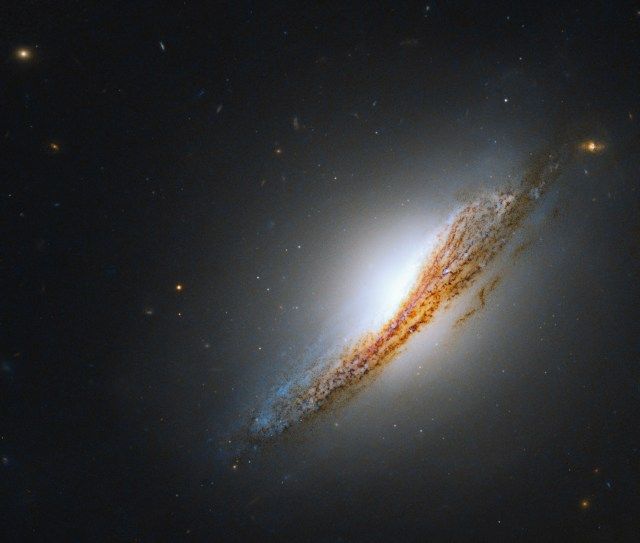 Black background dotted with galaxies. A large, nearly edge-on galaxy fills the right side of the frame. Its dusty, orangish-brown silhouette extends from center-bottom to the upper-right while its bright-white, lens-shaped glow extends above and below its dust lane.