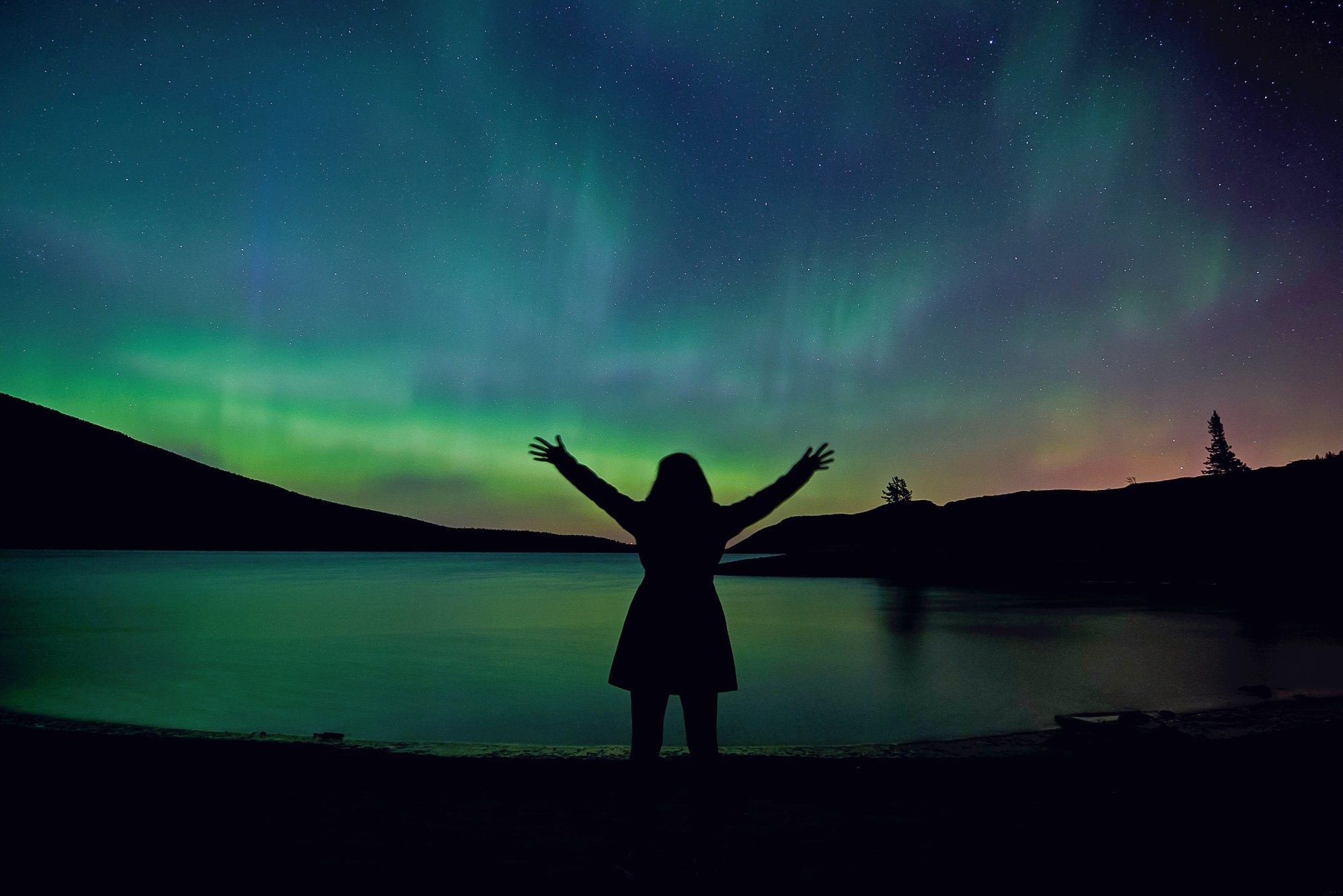 A woman reaches joyfully toward an aurora-lit night sky, her fingers spread wide. Green light shines above the horizon on the left, and orange light above the horizon on the right. over her head the sky has dancing green streaks. All the light is reflected in a lake at her feet.