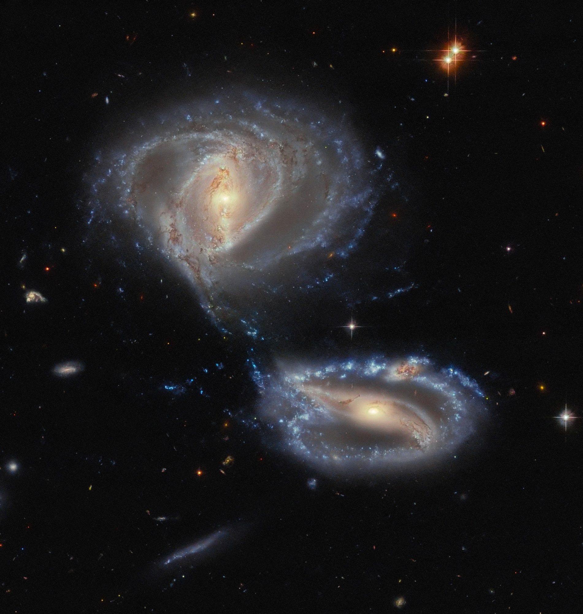 Two spiral galaxies. Each glows brightly in the center, where a bar stretches from side to side. The upper one is rounder, and its arms form two thin rings. The lower galaxy is flatter and its arms make one outer ring; a dusty knot atop its upper arm marks out a third object. Gravity is pulling gas and dust together where the galaxies come close. A number of small galaxies surround them on a black background.