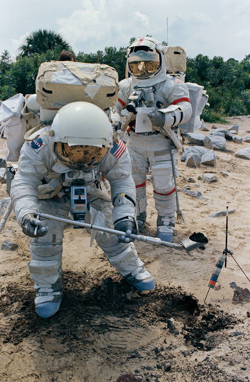 Two astronauts in spacesuits carry gear for digging in the soil in Florida.