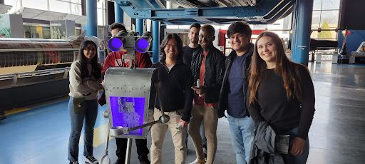 Seven people in their late teens/ early twenties wearing casual clothes stand in a well-lit, industrial space and look at the camera, smiling. A robot poses with them, also looking at the camera. One of the people is mostly blocked by a robot’s two round blue “eyes” that are affixed on either side of a grey box (the “head). The robot’s head and eyes are attached by a thin metal rod to a body that looks like the side-on view of an extra tall mailbox. The mailbox body tapers down to a smaller waist. The robot’s “arms” are visible, the right one down at it’s side and the left one raised so it’s “hand” - a metal ring, is visible in front of its body. 