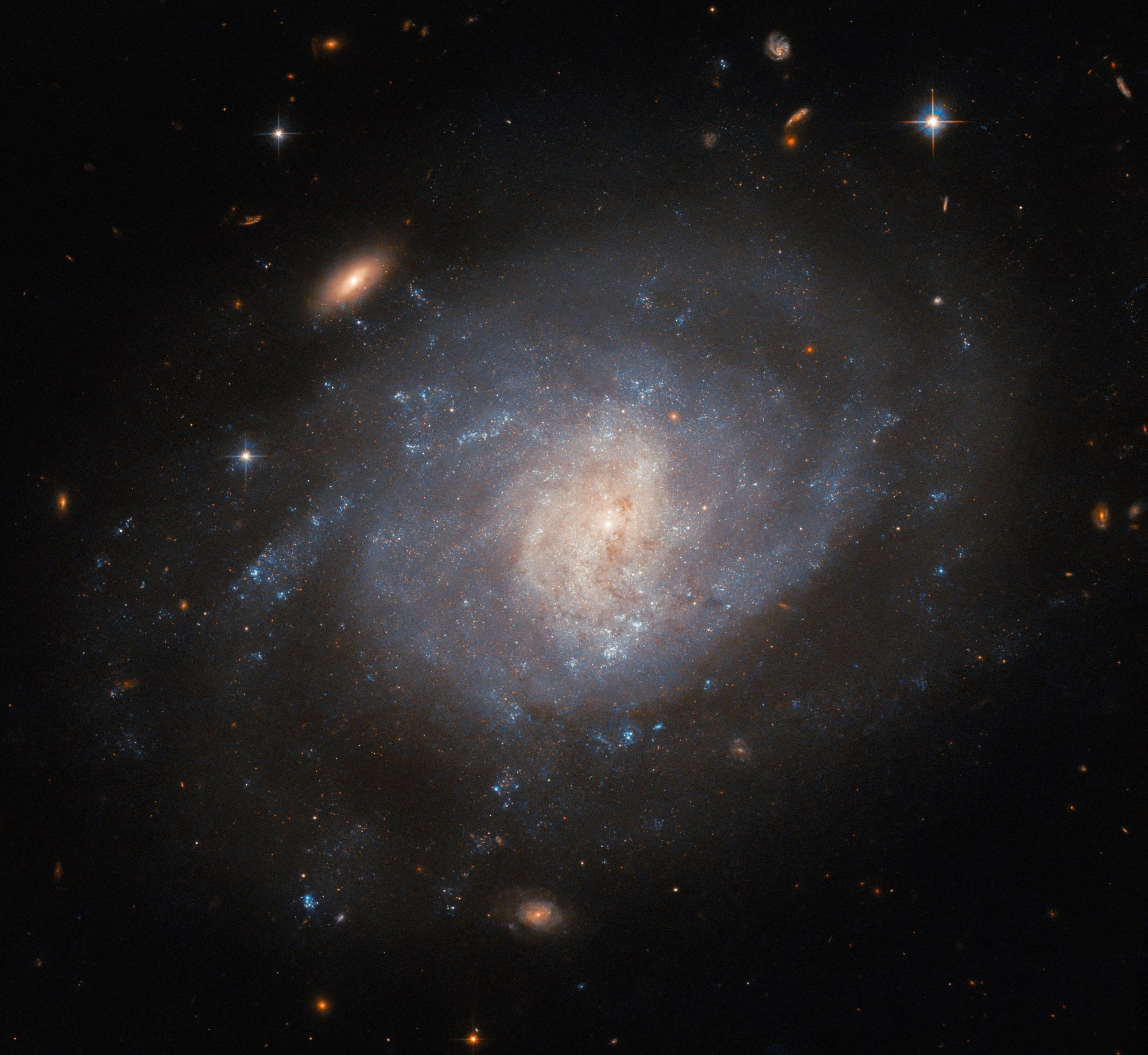 A spiral galaxy, seen face-on from Earth. The spiral arms of the galaxy are bright but not well defined, merging into a swirling disk with a faint halo of dimmer gas around it. The core glows brightly in a lighter color and has a bit of faint dust crossing it. Two redder, visually smaller galaxies and a bright star are prominent around the galaxy, with more tiny objects in the background.