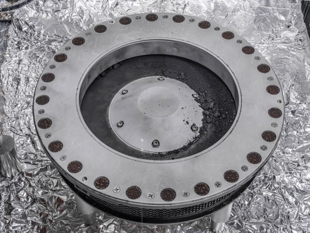 Silver, circular sample head with black and dark gray rocks and dust sample inside