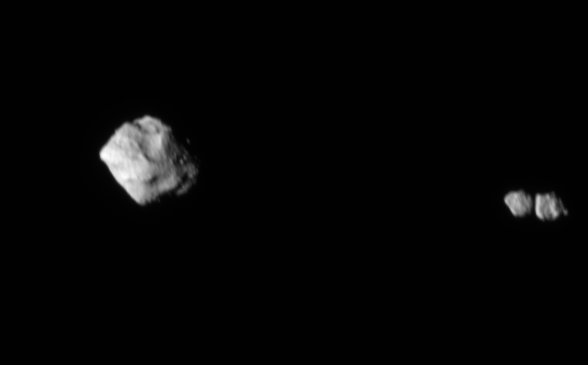 Against the blackness of space, a diamond-shaped asteroid taking up about a third of the height of the image is on the left-hand side. It's lit from the left. At the very right of the image, two small rocks are next to each other. There's a black line in between them which is a shadow from the left-hand rock. 