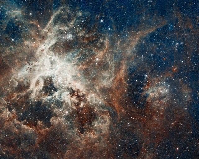 Tones of rusty-brown, blue, white, and a smattering of red fill the scene. Left of center holds bright-white tendrils of gas and a star cluster. Surrounding the cluster are tendrils and clumps of rusty-brown gas. Upper-left background glows faintly dark blue, while the lower half of the image holds a black background. Many stars dot the scene, their light shining within or through the nebula.