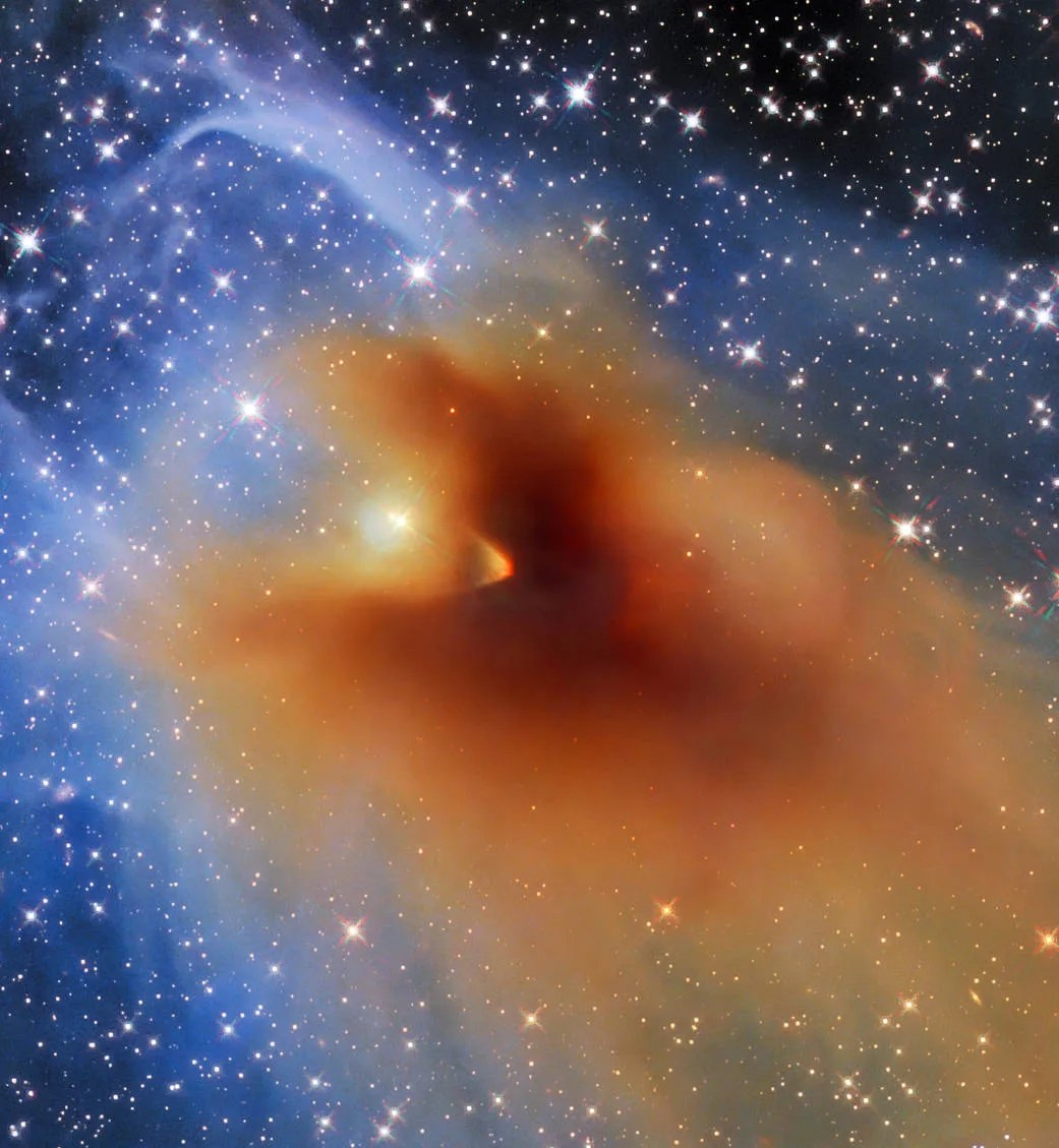 The image shows an irregularly-shaped bright orange object composed of dense gas and dust, which appears darker and more compact at the centre. This dense cloud, called CB 130-3, is outlined by thinner gas and dust in light shades of blue. The background shows a multitude of bright stars against a black background.