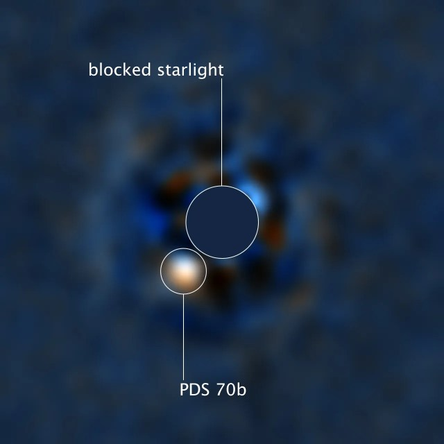 Blue background. Center of image is a disk blocking the light of a star. Below and just to the left of the disk, at about seven o'clock, is a bright white point. This is PDS 70b.