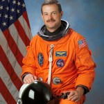 Photo of astronaut Scott Altman in his orange spacesuit. American flag is behind him.