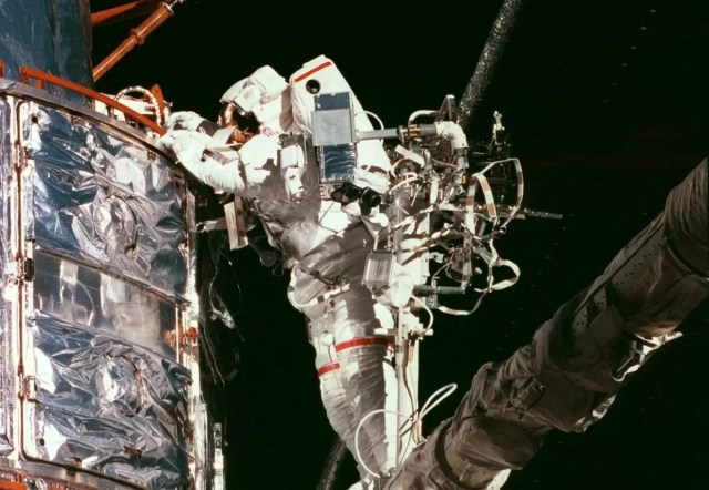 Astronaut standing on the extended robotic arm. On the left there is Hubble, which the astronaut is touching as part of the repair.