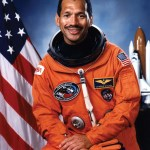 Photo of astronaut Charles Bolden in his orange spacesuit. American flag is behind him.