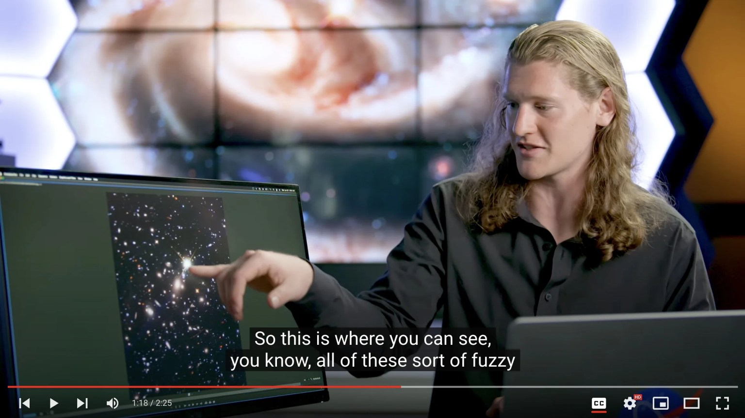 Scientist sits and points to a screen with a Hubble image while he discusses the science.