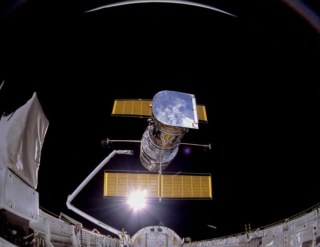 Image taken of the 1990 deployment of the Hubble Space Telescope. Hubble, bright and silver, reflects the Earth below, on either side of Hubble there are two golden solar arrays. At the bottom of the picture you can see the body of the Space Shuttle Discovery as well as the grapple arm letting go of Hubble.
