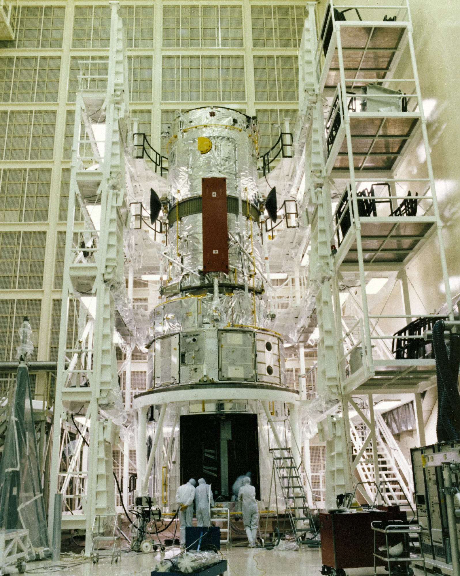 A partially constructed Hubble telescope is surrounded by scaffolding in a clean room. Technicians peer into a dark square opening at its base.