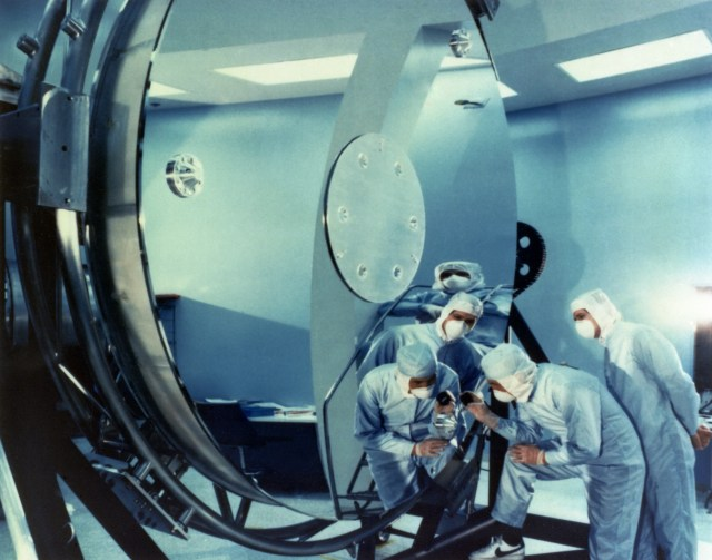 Technicians in blue suits and masks cluster around and are reflected in Hubble’s primary mirror while they inspect it.
