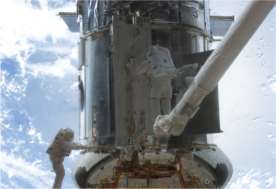 Two astronauts working on Hubble while in space. One astronaut is on the left, holding on to the lower handrail of Hubble. The astronaut on the right is standing on top of the CANADARM, a long robotic arm, he works on the center bottom of Hubble. In the distance behind Hubble, the blue Earth is visible with wisps of white clouds.