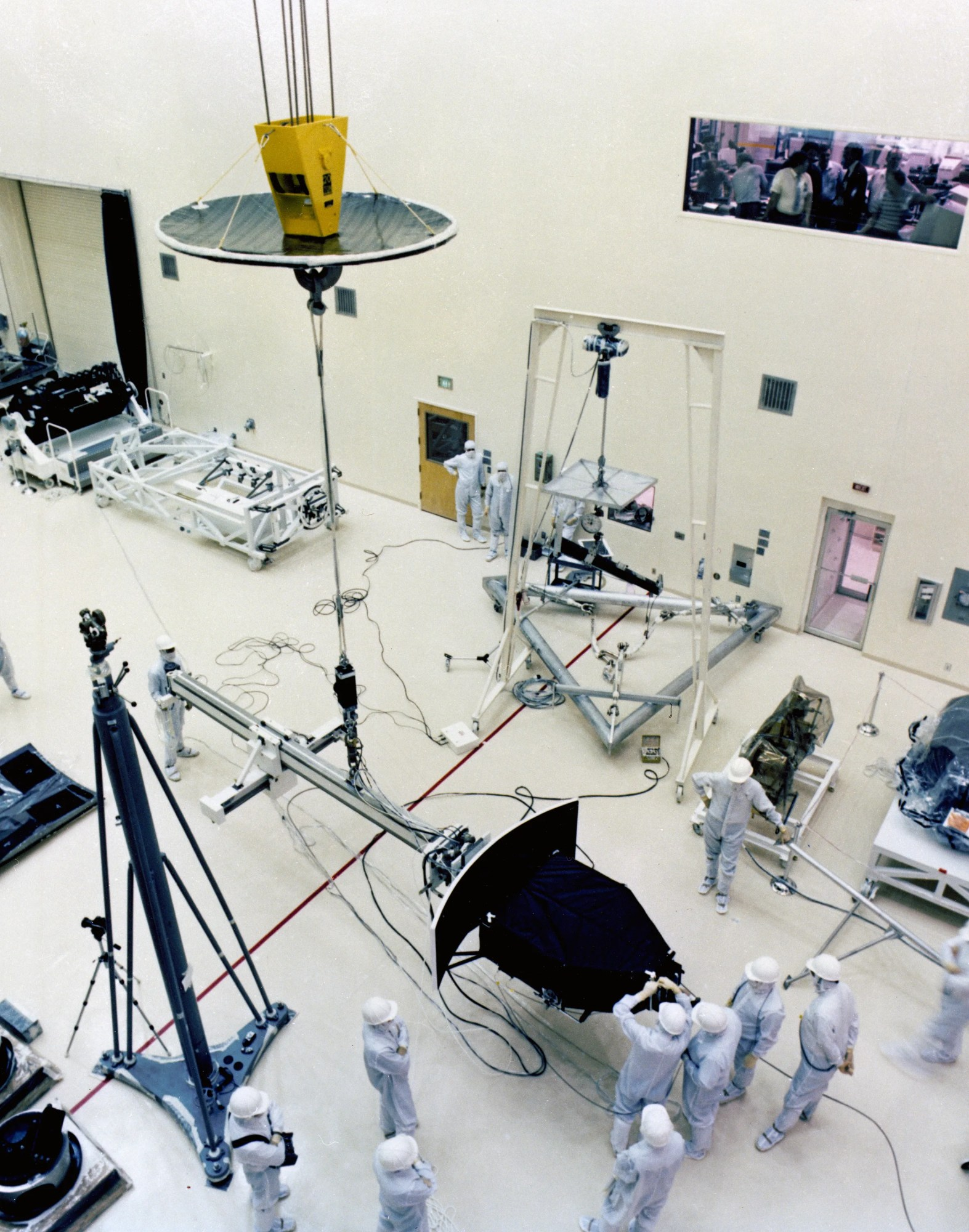 A group of white-clad and helmeted workers are seen from above as they work on Hubble’s Wide Field and Planetary Camera in a clean room.