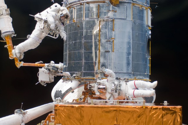 Two astronauts service the Hubble telescope. The one on the left is attached to a robotic arm. The one in the center waves to the camera.