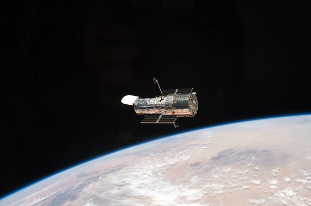 Image of Hubble seen floating above the Earth in orbit. Under Hubble, the sandy colored landmass is visible through wisps of clouds. On the upper half of the image, the darkness of space is seen.