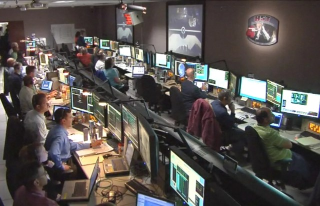 Photograph of a group of engineers sitting in front of their computers inside of the "Space Telescope Operations Control Center." This image was taken during Hubble's Servicing Mission 4, which is on the screen in the front of the room.