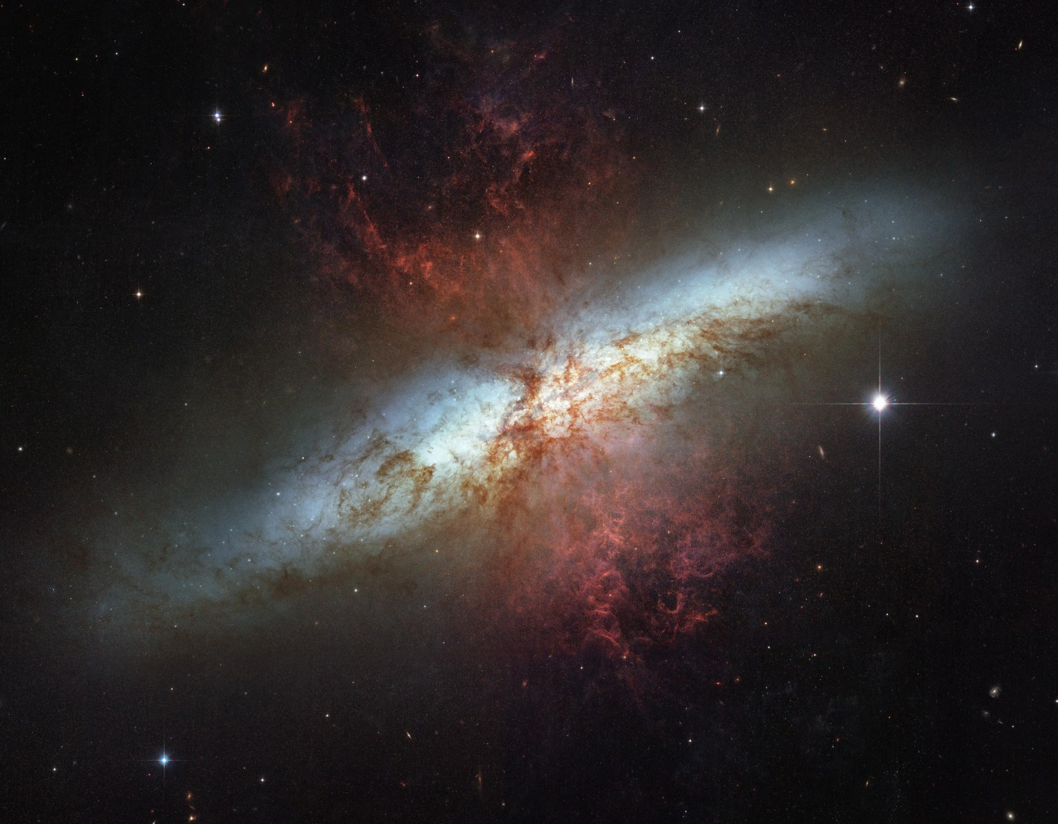 A white band of stars that cuts across a black background, from the lower left to the upper right, is the galaxy M82. Reddish brown gas and dust overlays the galaxy concentrated in the center of the image and fanning out above and below the white band of the galaxy. Black background is dotted with stars.