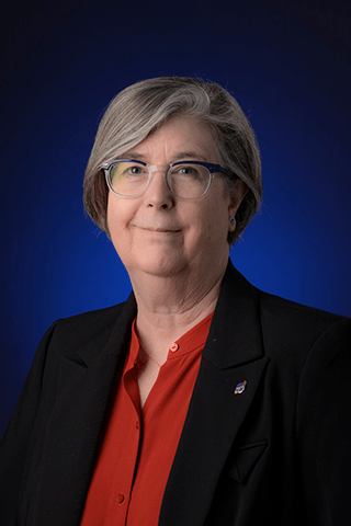 Portrait photo of a woman with short grey hair wearing a red shirt and dark blazer in front of a dark blue background
