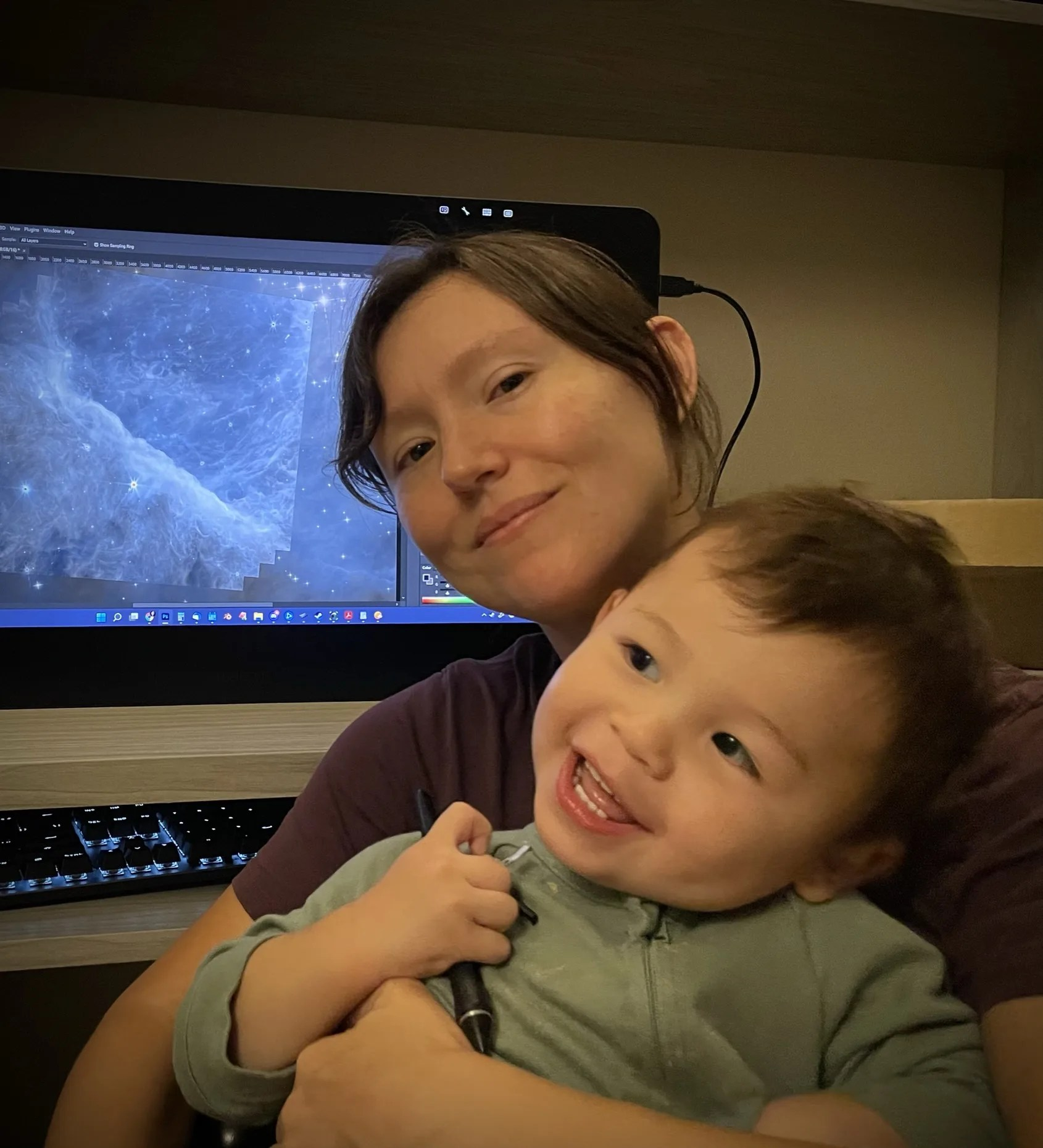 Photo of a smiling woman holding a smiling toddler, a computer screen displaying James Webb Space Telescope imagery is in the background.