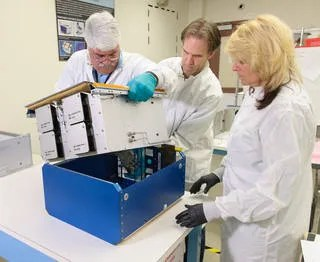 Two male and one female scientist assembled removing a white piece of equipment from a blue casing.