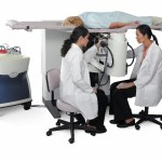 Two medical workers in white coats work at an imaging machine. A patient in a hospital gown lies on a platform on top of the machine.