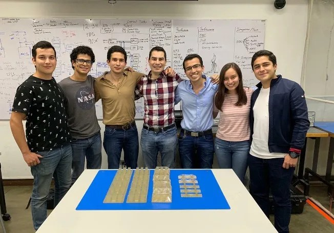 Group of 6 men and 1 woman standing with their arms around each other; a whiteboard filled with writing is in the background and a blue mat with small clear components sits on a table in front.