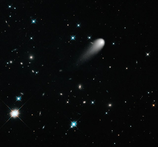 Black background dotted with stars and galaxies. The bright-white comet is in the upper right quadrant of the image, just to the upper right of center. Its tail is pointing  toward the lower left corner. 