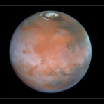 Rusty-red Mars with a haze of white clouds and a white north polar cap.